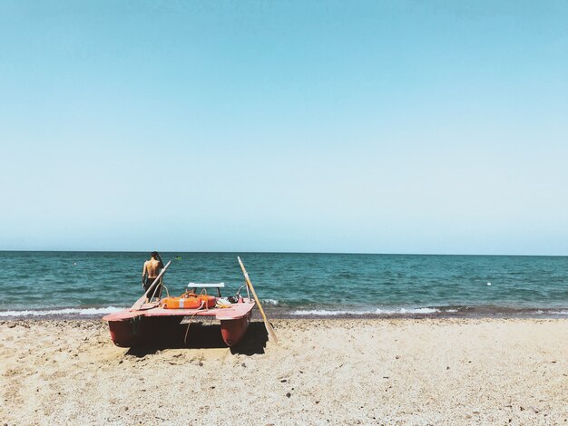 Person standing near a boat on the beach shore with a blue sky