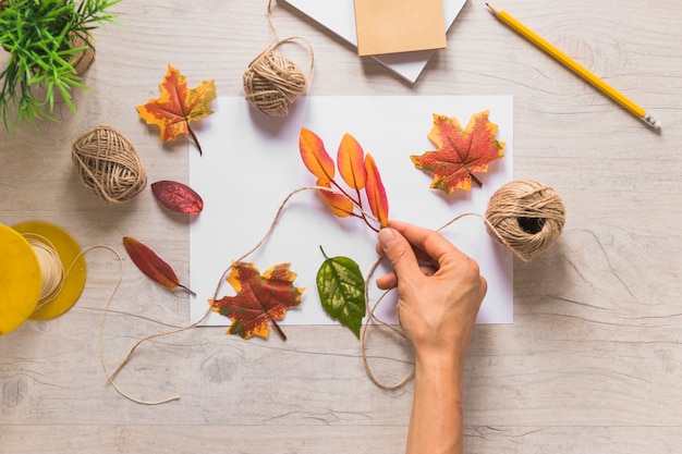 A person tying fake leaves with string spool on white paper over the wooden textured background