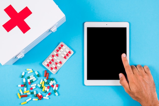 A person using digital tablet with first aid kit and capsules on blue background