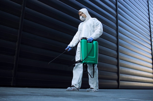 Person in white chemical protection suit doing disinfection of public areas to stop spreading highly contagious corona virus