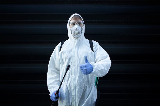 Человек в белом костюме химической защиты держит распылитель с дезинфицирующими химикатами, чтобы остановить распространение очень заразного вируса