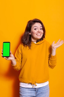 ディスプレイスマートフォンの垂直緑色の画面を持つ人。スタジオのオレンジ色の背景の上にスマートフォンのクロマキーで孤立したモックアップテンプレートを示すポジティブな大人。電話を使用している女性