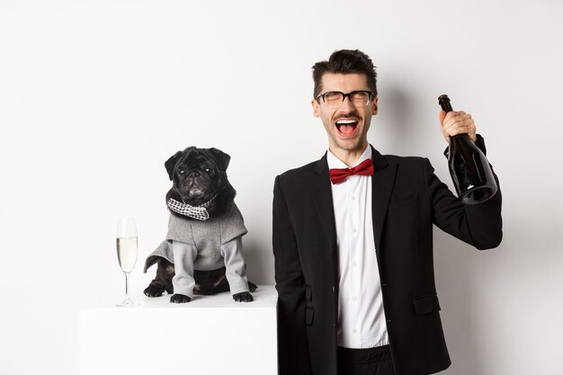 Pets, winter holidays and New Year concept. Happy man celebrating Christmas party pet, standing with cute dog in costume, drinking champagne and rejoicing, white background