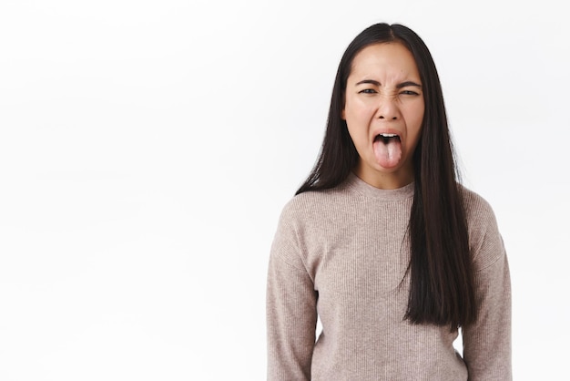 嫌な味が嫌い嫌いな若いアジアの女の子は嫌悪感を感じてピクルスを試み、悪臭を吐きたいようにスティック舌を細めるのが嫌い失望を表現する白い背景