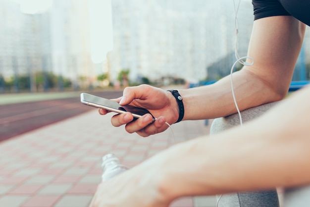 Phone in muscular hand of guy sitting in the city in the morning. He holds bottle of water and headphones.