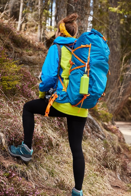 Фотография активной туристки, которая поднимается на холм гор, несет большой рюкзак, носит ботинки.