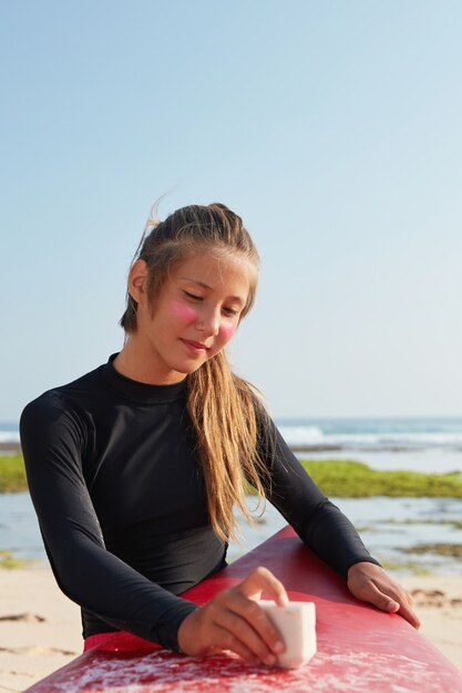 잠수복을 입은 활동적인 젊은 웨이크 서퍼의 사진, 조랑말 꼬리, 왁스 사용, 바위 해안선 근처에서 포즈, 비버 꼬리 착용