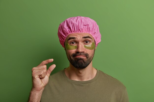Photo of adult man with hydrogel patches under eyes for reducing puffines, wears waterproof shower cap, makes small gesture, tells he needs not much time for beauty treatments and date preparation