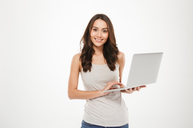 Photo of affable woman with long brown hair holding silver personal computer posing on camera, isolated over white wall