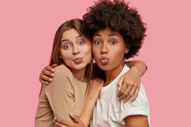 Photo of affectionate lovely mixed race women stand closely to each other, make kiss grimace