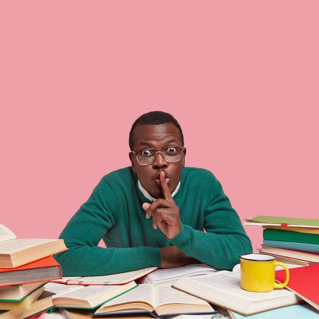 Photo of African American male bachelor keeps fore finger on lips, asks not making noise while studying, wears green sweater