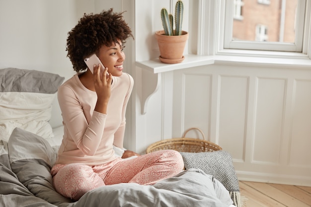 Photo of Afro American lady makes appointment via cellular, dressed in casual clothes, sits in lotus pose on bed, wears casual clothes, enjoys conversation with friends, has day off, poses in bedroom