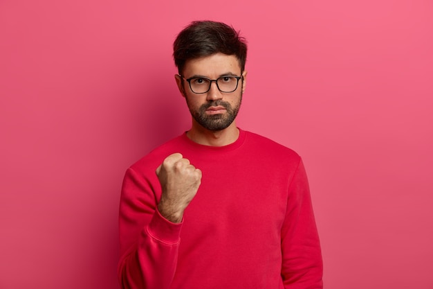 Photo of aggressive confident adult man has dark hair and beard, clenches fist and looks seriously , doesnt afford being insulted, shows his power, wears spectacles and red sweater.