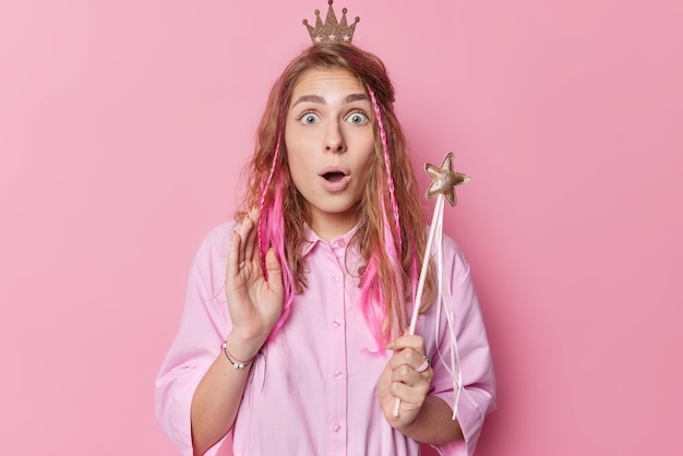 Photo of amazed long haired young woman stares bugged eyes holds breath keeps mouth opened cannot believe in shocking news holds magic star wand wears crown poses against pink studio background.