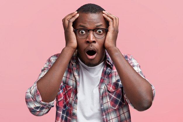 Photo of amazed shocked African American male looks in terror and with eyes popped out, keeps hands on head, keeps mouth widely opened, expresses unexpectedness and surprisment, isolated on pink wall