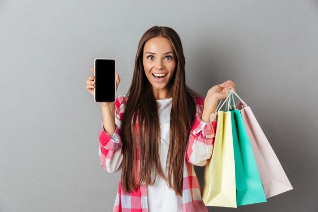 Photo of amazed young brunette holding shopping bags, showing blank mobile screen