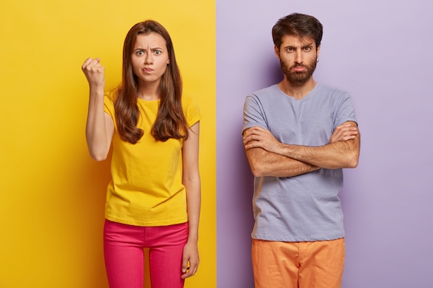 Free photo photo of angry annoyed woman raises clenched fist, expresses negative emotions, wears yellow casual t shirt and pink trousers, sad man keeps arms folded, feels offended, being abused by someone