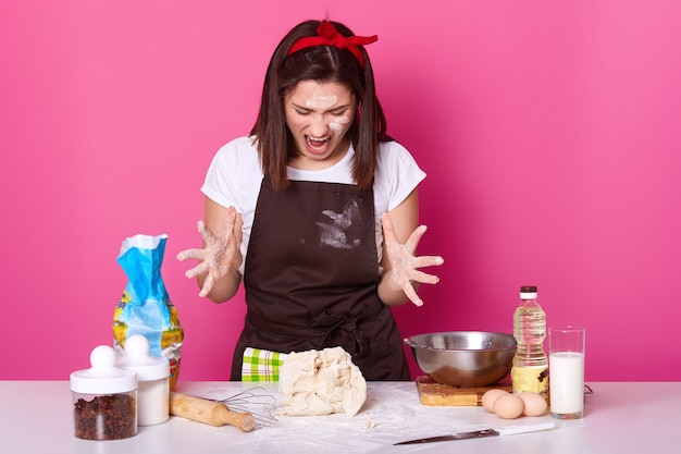 Photo of angry brunette baker being sick and tired of kneading dough, dressed in casual t shirt and brown apron dirty with flour. female spreads her fingers while shouting something. food concept.