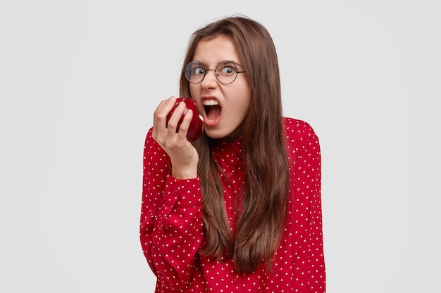 Photo of angry hungry young woman bites apple with annoyance, being in bad mood as keeps to diet, wants to eat