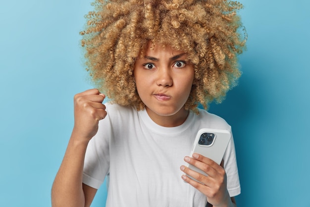 Фотография сердитой возмущенной кудрявой женщины с кошельками, сжимающей губы в кулак, сердито смотрит и использует современный мобильный телефон для отправки текстовых сообщений, одетая в повседневную белую футболку, изолированную на синем фоне