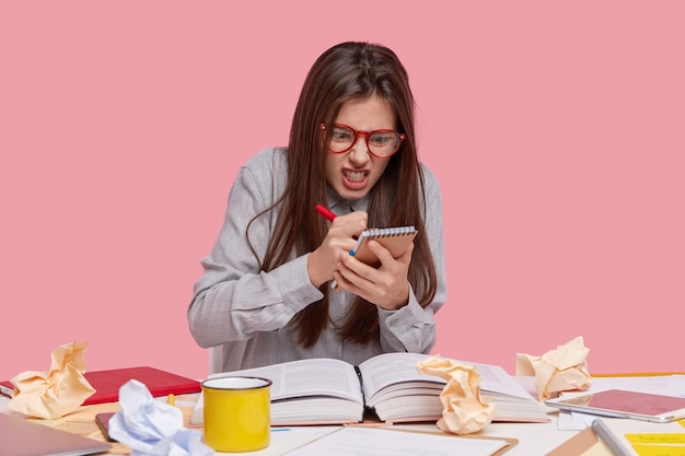 Free photo photo of angry outraged young female author records information with irritated expression, clenches teeth from anger, holds pen and spiral notebook