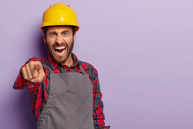 Photo of annoyed foreman points, yells with reproach, shouts angrily at collegue, wears yellow helmet, shirt and apron. Irritated builder solves problem at work