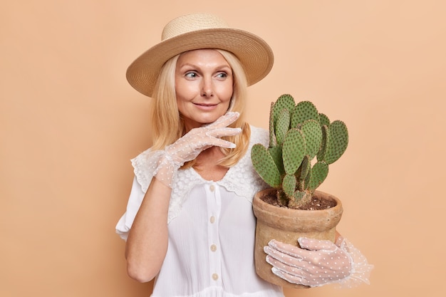 Foto di aristocratica signora di mezza età indossa cappello camicetta bianca e guanti di pizzo tocca il mento guarda delicatamente da parte tiene cactus in vaso dà consigli su come prendersi cura della pianta d'appartamento isolata sul muro beige