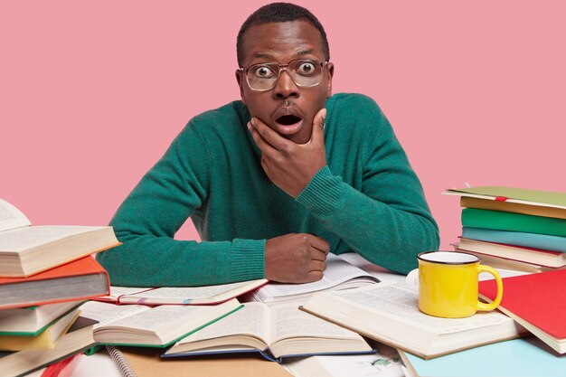 Photo of astonished black young man holds chin, stares in disbelief, has many opened books around table