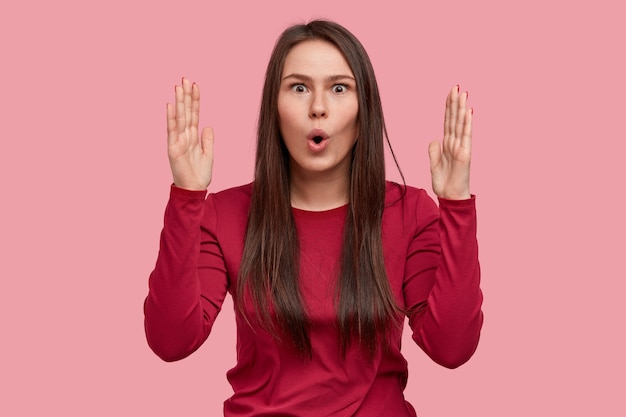 Photo of astonished brunette lady shows something big or huge with both hands, has surprised facial expression, wears red outfit