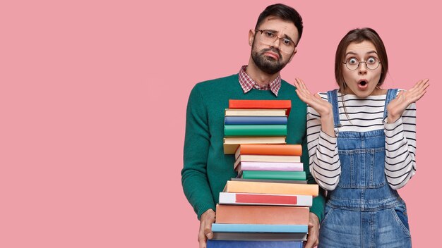 Photo of astonished Caucasian young lady in denim overalls, tired unshaven male carries textbooks