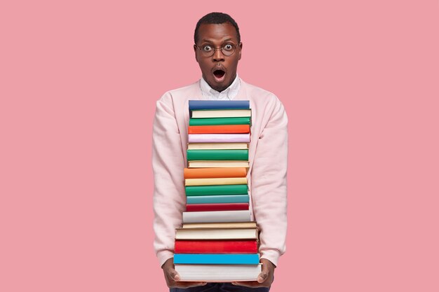 Photo of astonished handsome black young man carries many books, keeps jaw dropped, wears casual clothes, amazed by difficult tasks