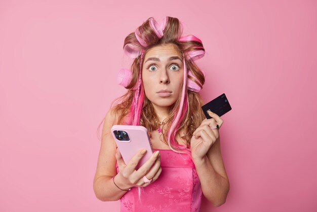 Photo of astonished woman stares bugged eyes cannot believe in shocking news makes payment online holds credit card and cellular wears hair curlers stylish dress isolated over pink background