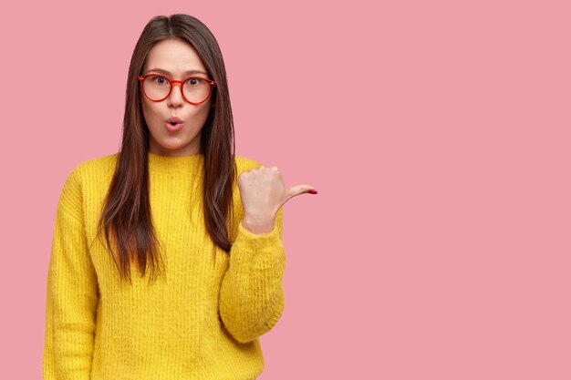 Photo of astonished young woman opens mouth with amazement, points right, reacts to awesome thing happend aside, wears oversized sweater