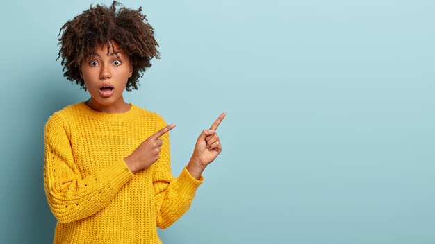 Photo of astound good looking woman shouts from shocking news, points on right empty corner, wears yellow jumper, surprised by unexpected price, shows something. Copy space for announcement