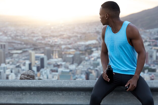 Photo of athletic young jogger with muscular body, dark skin, turns back, admires beautiful panoramic view, wears blue casual vest, feels thirsty after long running race, has lack of energy.