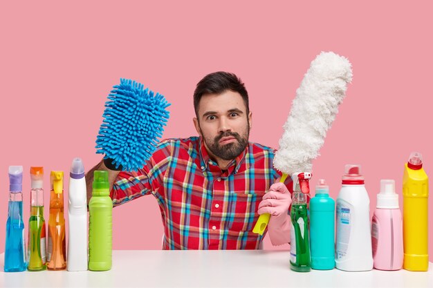 Photo of attentive young male janitor looks scrupulously, holds sponge and dust brush, wears red checkered shirt