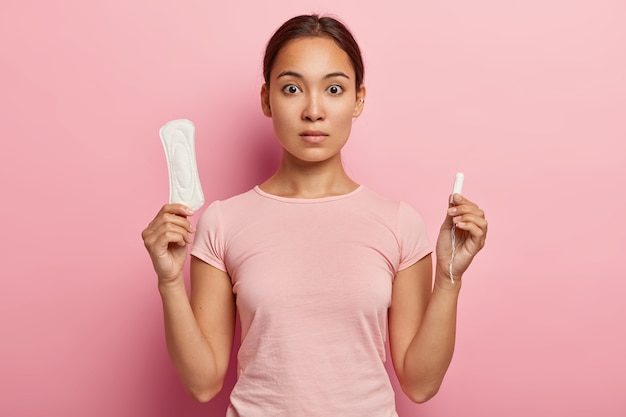 Photo of attractive Asian woman holds cotton tampon and sanitary napkin, has surprised look, monthly menstrual cycle, wears pink t shirt. Protective care for womens health. Fertilization concept