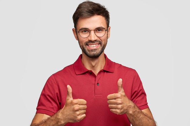 Photo of attractive bearded young man with cherful expression makes okay gesture with both hands, likes something, dressed in red casual t-shirt, poses against white wall, gestures indoor