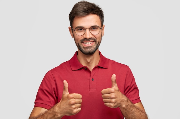 Photo of attractive bearded young man with cherful expression makes okay gesture with both hands, likes something, dressed in red casual t-shirt, poses against white wall, gestures indoor