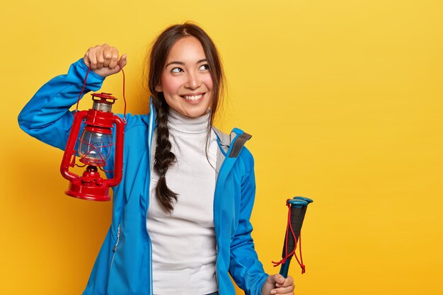 Photo of attractive brunette woman with pensive glad expression, holds red gas lamp, trekking poles, covers long distance on mountain path, wears turtleneck and blue jacket, recalls pleasant moment