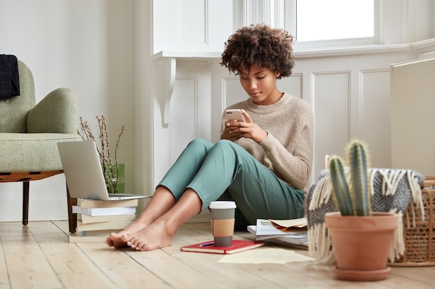 Photo of attractive busy woman has bushy hair, reads business data on cell phone, prepares for session