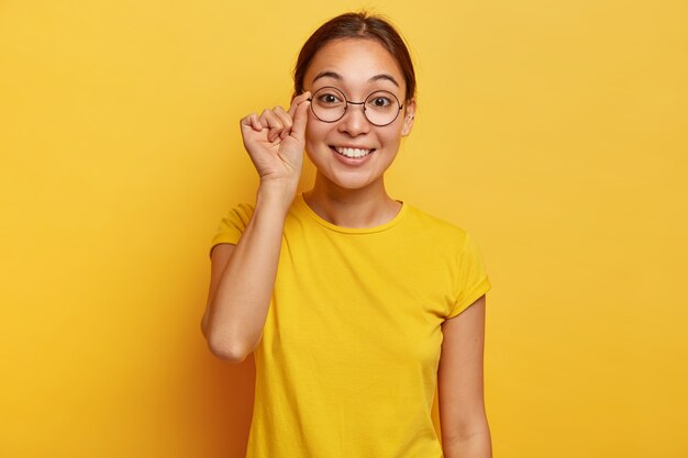 Photo of attractive female looks curiously, has happy expression, touches frame of spectacles, wears yellow t shirt, reads good news, concentrated , poses indoor. Human face expressions