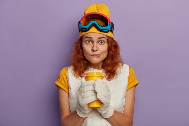 Photo of attractive female model has ginger hair, bites lips and holds takeaway cup with both hands, looks directly at camera wears yellow t shirt with vest