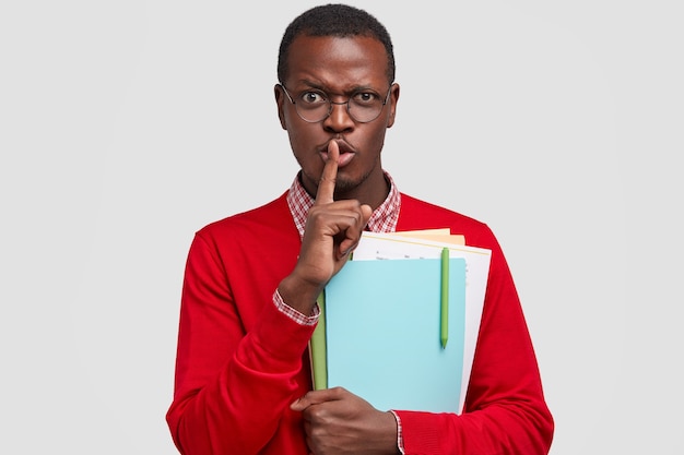 Photo of attractive secret man keeps index finger, demonstrates hush gesture, carries papers with pen