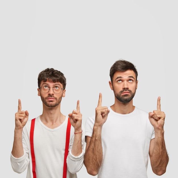 Free photo photo of attractive two bearded young men with serious expressions, point with both index fingers upwards, show cool copy space, advertise something over white wall, dressed in casual clothing