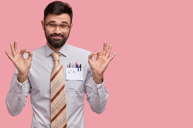 Photo of attractive unshaven man makes okay gesture with both hands, dressed in formal outfit