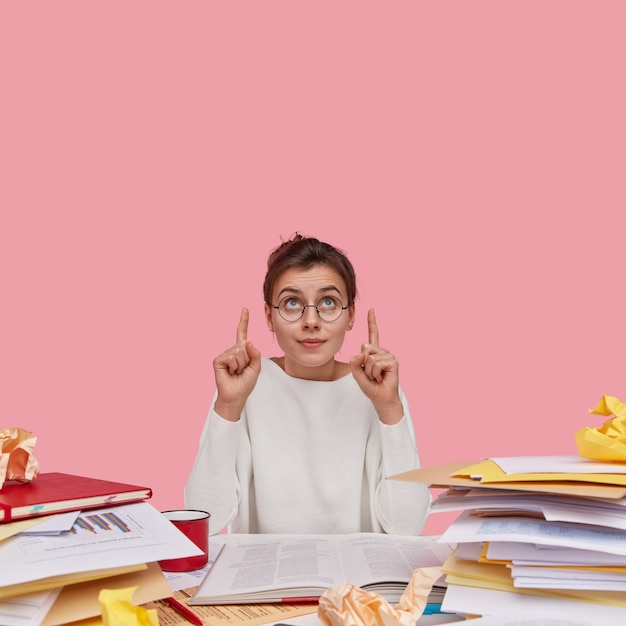 Free photo photo of attractive woman has dark hair, dressed in white sweater, points with both fore fingers upwards, sits at desktop