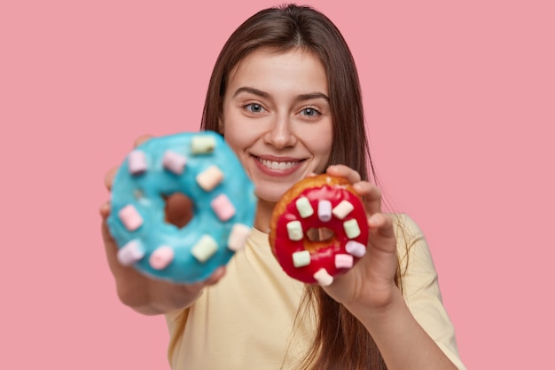 Foto gratuita la foto di una donna attraente tiene gustose ciambelle, suggerisce di assaggiare, sorride positivamente, ha un aspetto accattivante