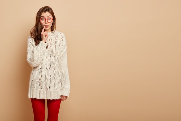 На фото привлекательная женщина держит указательный палец возле сложенных губ, смотрит с любопытством, удивленное выражение лица, слушает интригующую информацию, носит повседневный зимний свитер, стоит над бежевой стеной.