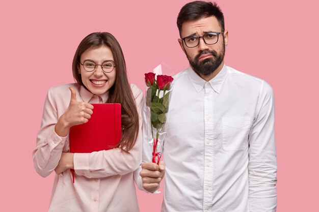 Фотография привлекательной женщины показывает жест одобрения, держит большой палец вверх, несет красный блокнот, озадаченный небритый мужчина держит букет роз